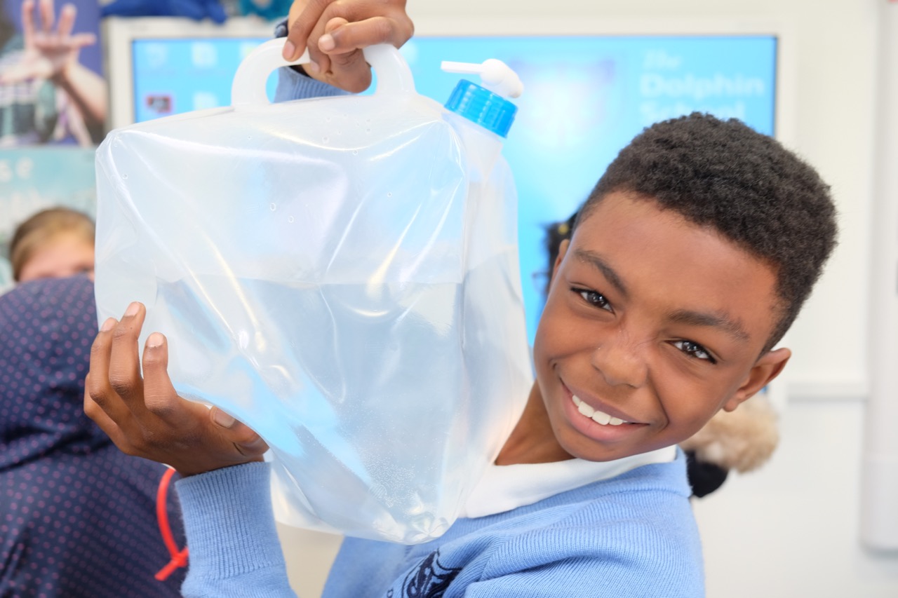 young boy in UK school uniform, smiling, holds up jerry can filled with water