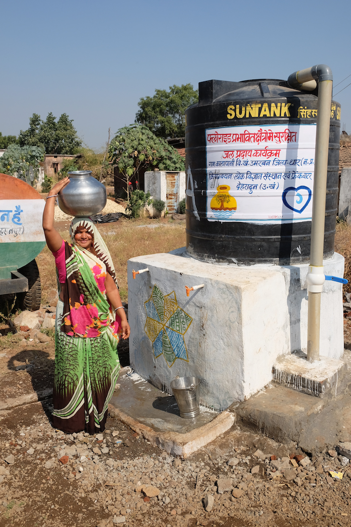Woman stands next to safe water tank with water container on her head